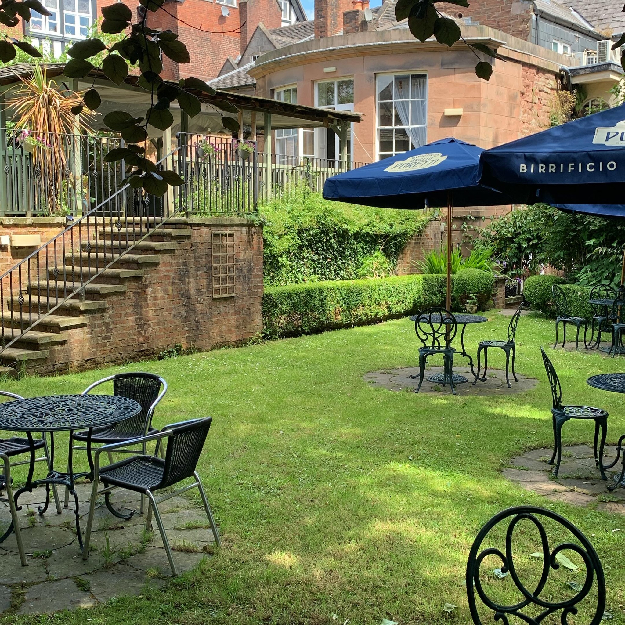 The Silk House beer garden lawn with bistro tables and umbrellas among mature trees and hedging